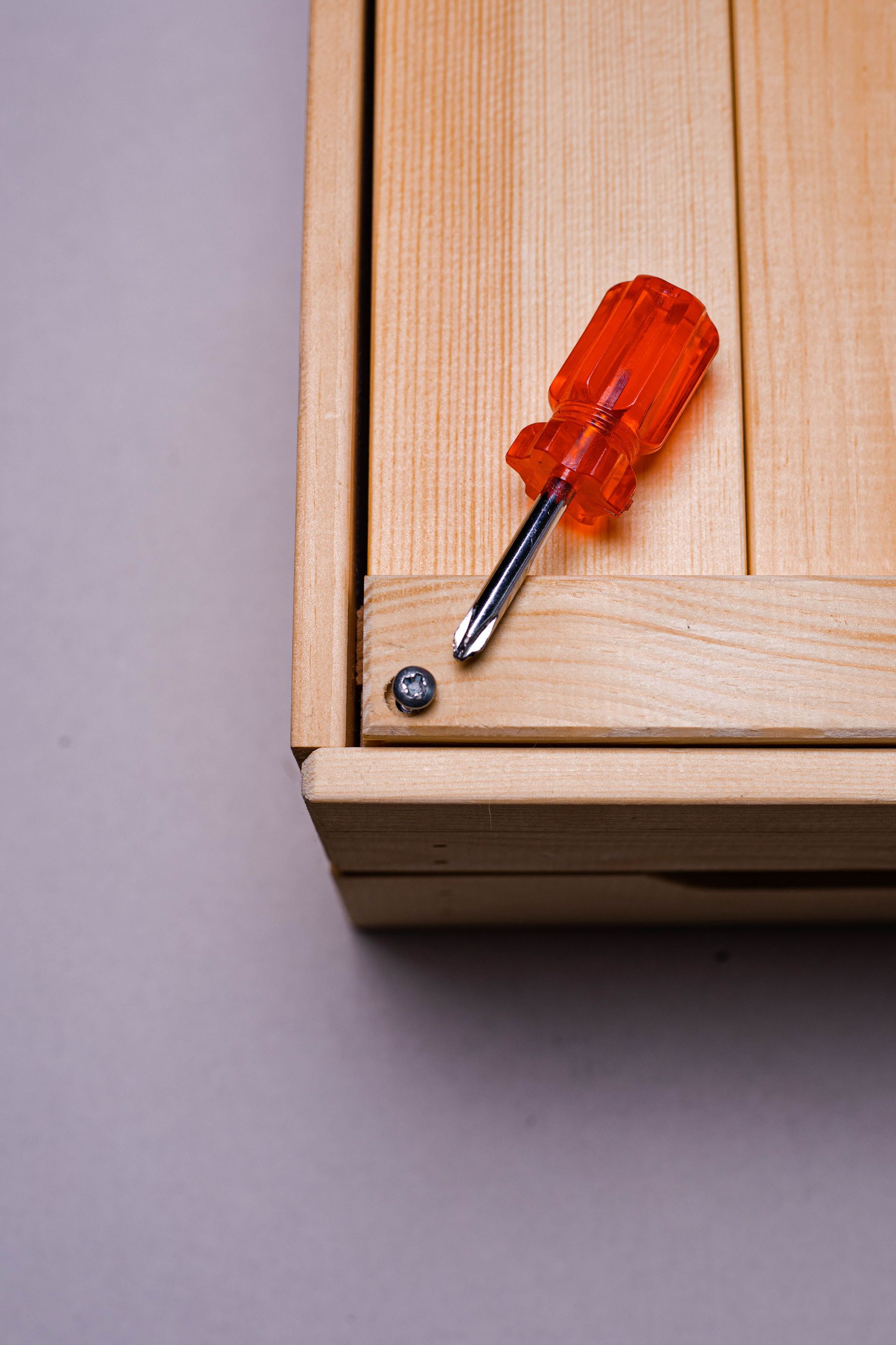 Close-up of a wooden box corner with an orange screwdriver on a grey surface.